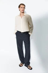 Colin Otto poses in a cream striped linen shirt, dark navy trousers, and black flip-flops against a white studio background.
