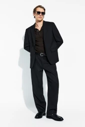 Colin Otto standing against a white backdrop wearing a black blazer, dark polo shirt, black trousers, and sunglasses.