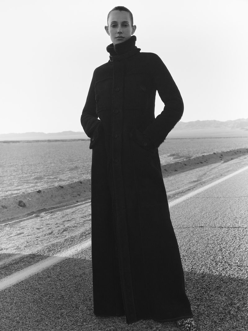 Woman in long, dark coat on desert road with distant mountains. Full-length portrait with muted, monochromatic color palette.