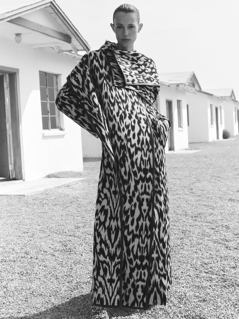 Fashion shot of a slender woman in a long black and white patterned coat, posing outdoors in front of white, one-story buildings on a gravel surface.