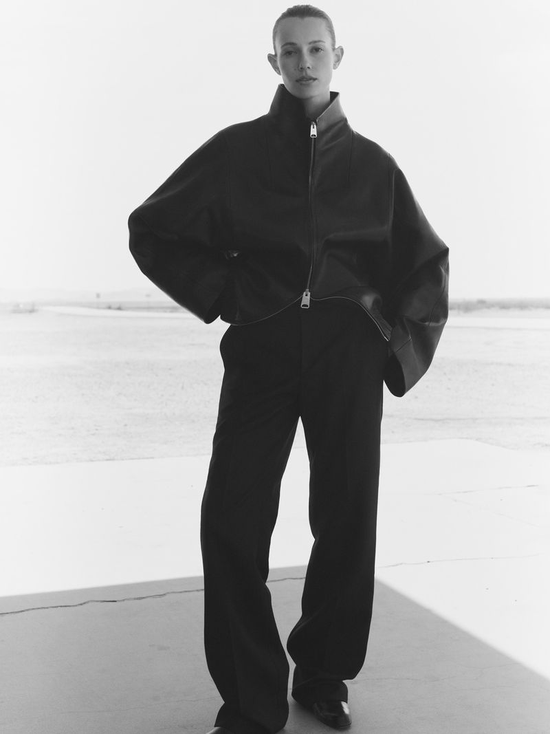 Monochrome fashion portrait of an adult woman posing in a leather jacket and wide-leg pants against a bright, neutral background.