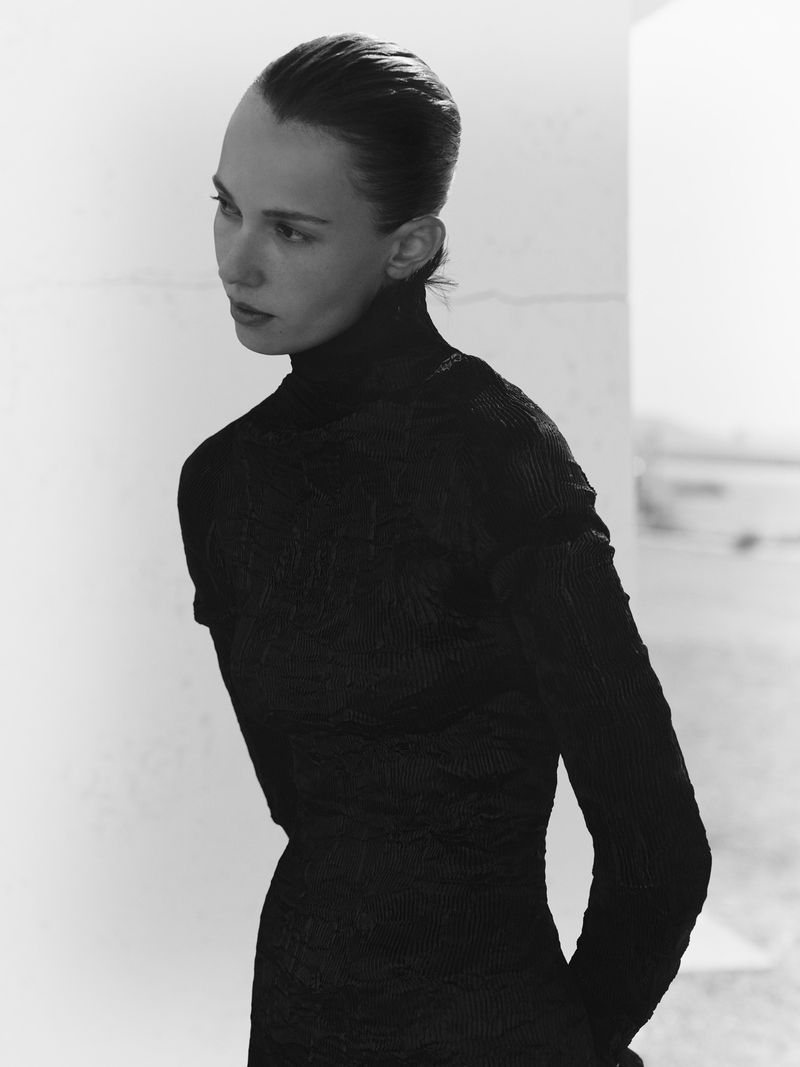 Portrait of a young woman in a black turtleneck dress, shot in black and white. The background is a blurred neutral color.
