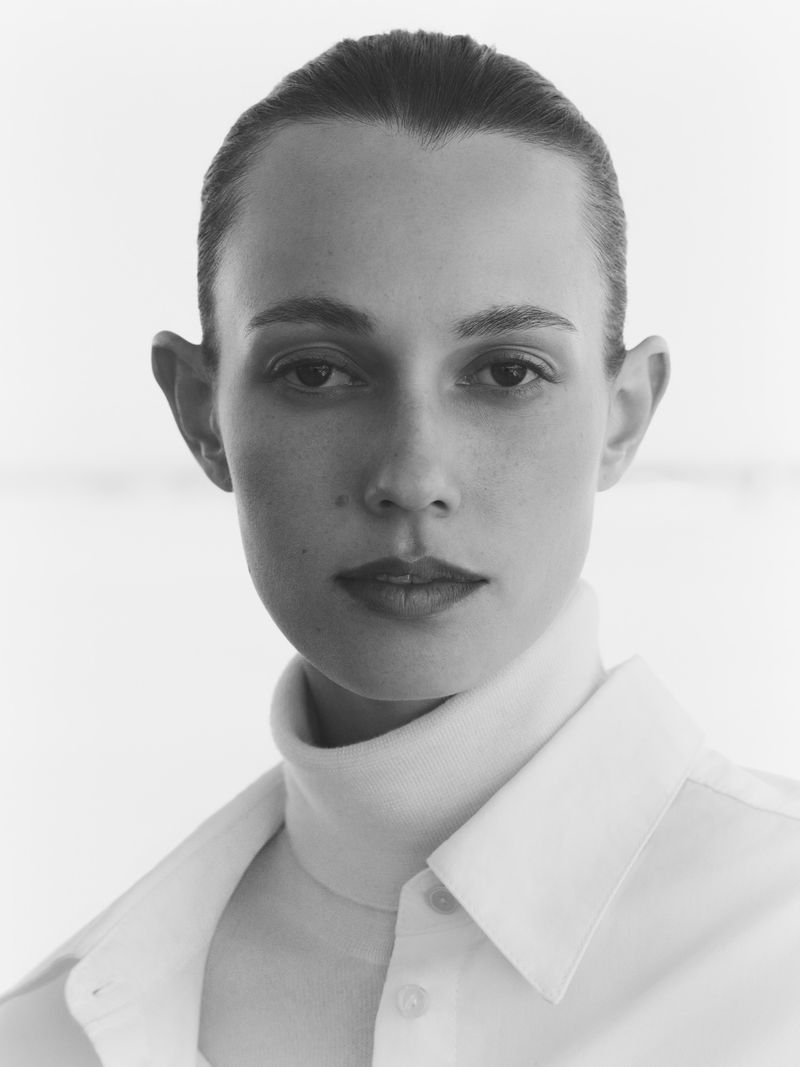 Monochrome portrait of a woman with dark hair and fair skin, wearing a white turtleneck and button-down shirt, against a bright backdrop.