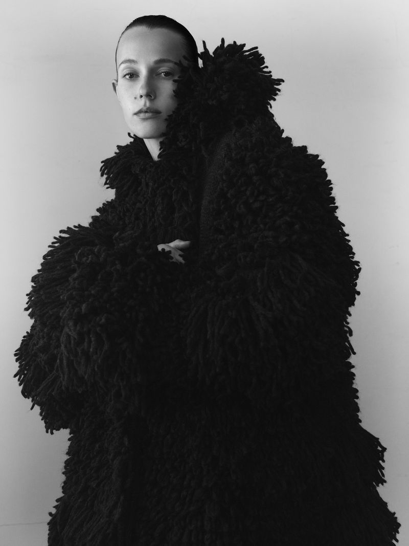 Portrait of a woman with bare head and black shaggy coat in studio, with minimal makeup and fair skin against a neutral background.