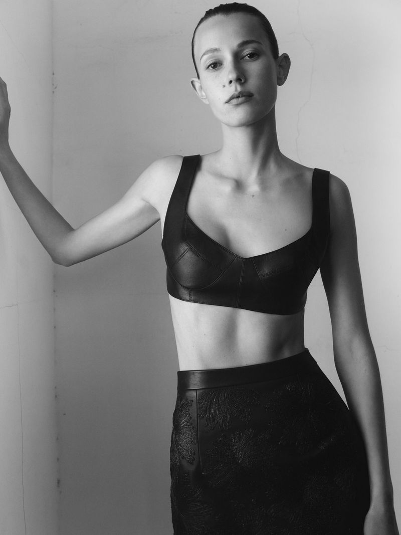 Monochrome portrait of a young woman in a bra top and skirt, indoor against a neutral wall in a studio setting.