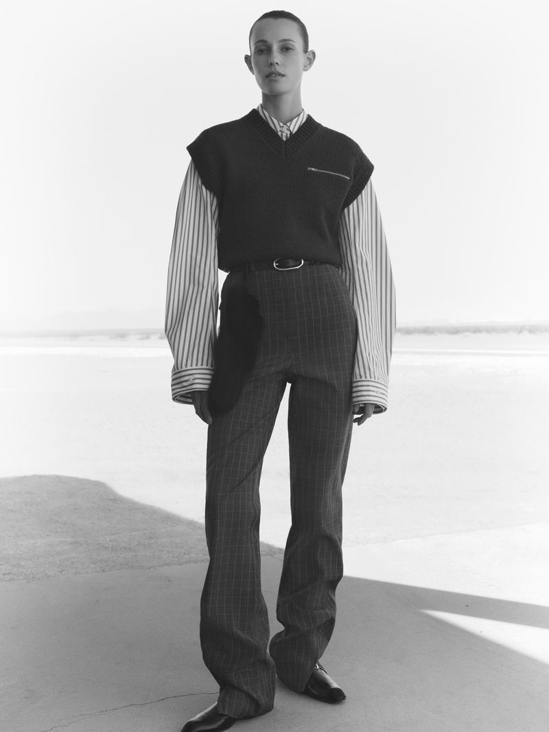 Monochrome full shot of a slim adult female model in pinstripe pants, striped shirt, and vest against a bright outdoor backdrop.
