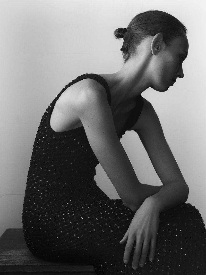 Black and white profile portrait of model Mali Koopman seated in a textured sleeveless black dress against a plain studio backdrop.