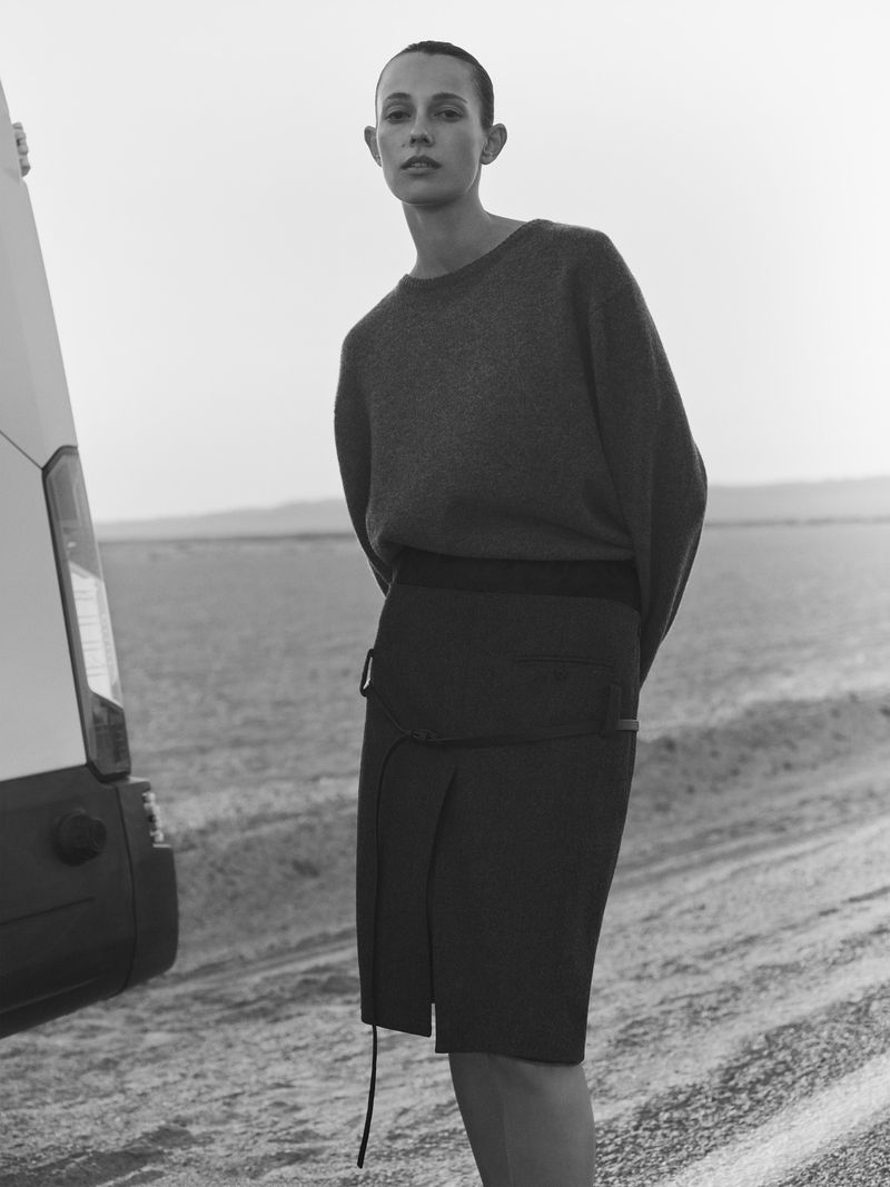 A slender adult woman with a buzzcut poses beside a white van on a desolate road, wearing a gray sweater and a dark midi skirt.