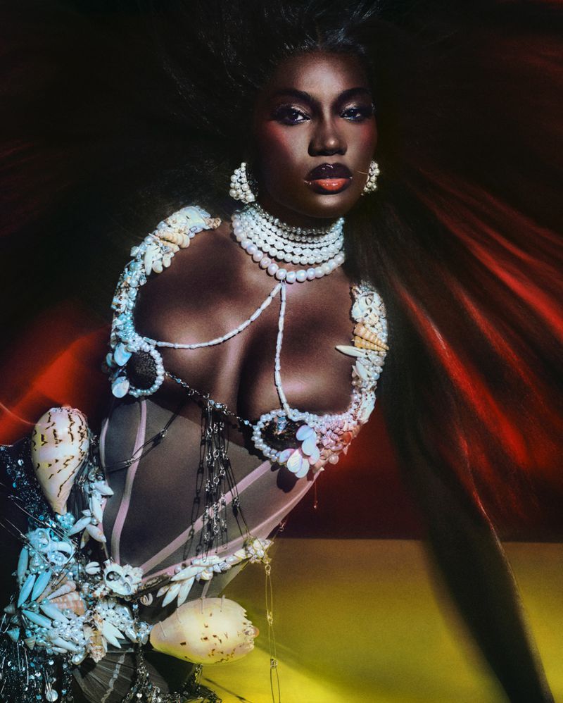 Dark-skinned woman with dramatic makeup, wearing a seashell-embellished bodice and pearl jewelry against a dark, dynamic background.