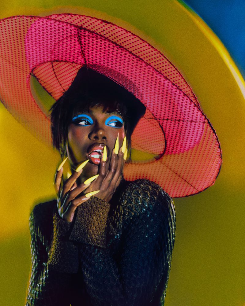 Glamour shot of a model with dark skin in a large pink hat with bright blue eye makeup and yellow fingernails, posing with hand to face.