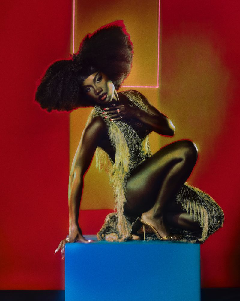Elegant Black woman with large afro hairstyle poses on a blue cube in a feathered dress against a red studio backdrop with neon square.
