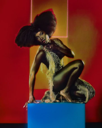 Elegant Black woman with large afro hairstyle poses on a blue cube in a feathered dress against a red studio backdrop with neon square.