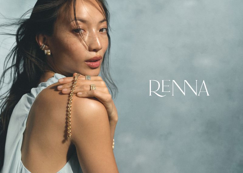 East Asian woman looking over her shoulder wearing gold ear cuffs, rings, and a draped beaded chain against a textured blue backdrop.