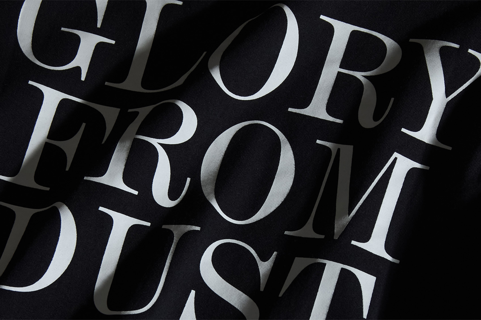 Close-up of black fabric with white typography reading GLORY FROM DUST, featuring soft folds and natural shadows.