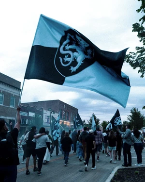 Crowd of supporters marching along a town street with a large black and light blue heraldic lion flag waving in the air.