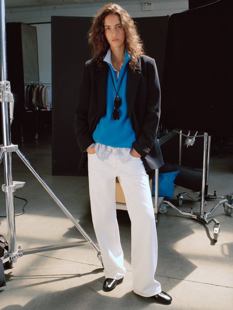Model wearing a black blazer, bright blue top, and wide-leg white trousers standing on a photography studio set.