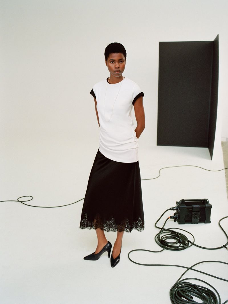 Model Victoria Fawole wearing a white top and black lace-hem midi skirt posing on a studio set with floor cables.