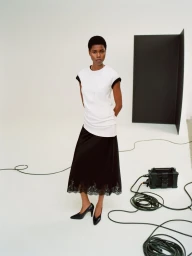 Model Victoria Fawole wearing a white top and black lace-hem midi skirt posing on a studio set with floor cables.