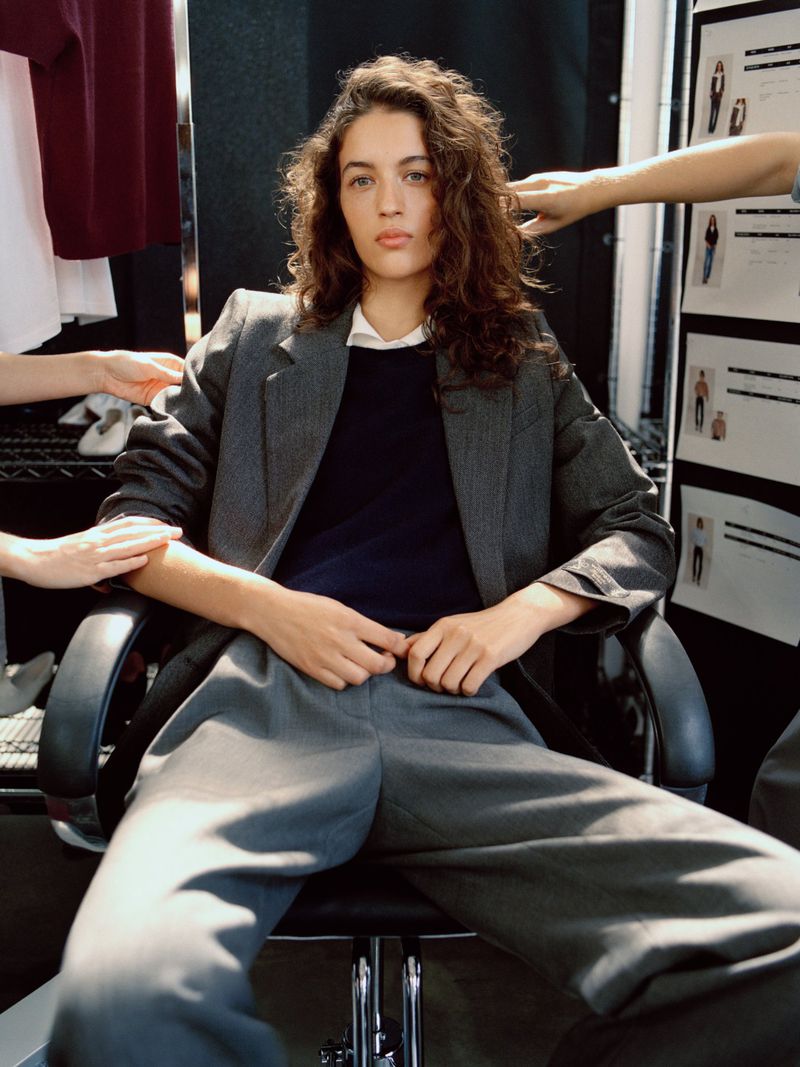 Model seated in a rolling chair wearing a grey tailored suit and navy sweater while crew hands adjust hair and clothing backstage.