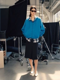 Model wearing an oversized blue zip hoodie, black skirt, sunglasses, and white heels in a photography studio with lighting gear.