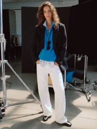 Model wearing a black blazer, bright blue top, and wide-leg white trousers standing on a photography studio set.