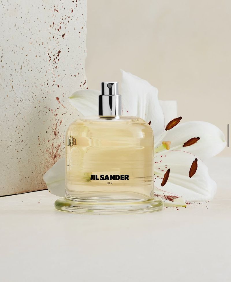 Jil Sander Lily perfume bottle in clear glass with pale golden liquid, placed beside a white lily flower and a speckled stone slab.