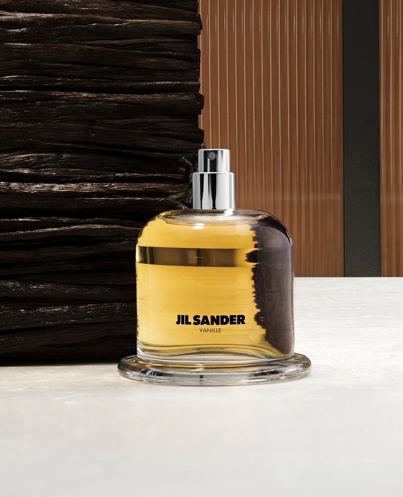 Jil Sander Vanille perfume bottle filled with golden amber liquid resting on a white stone surface beside dark textured vanilla pods.