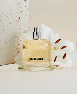 Jil Sander Lily perfume bottle in clear glass with pale golden liquid, placed beside a white lily flower and a speckled stone slab.