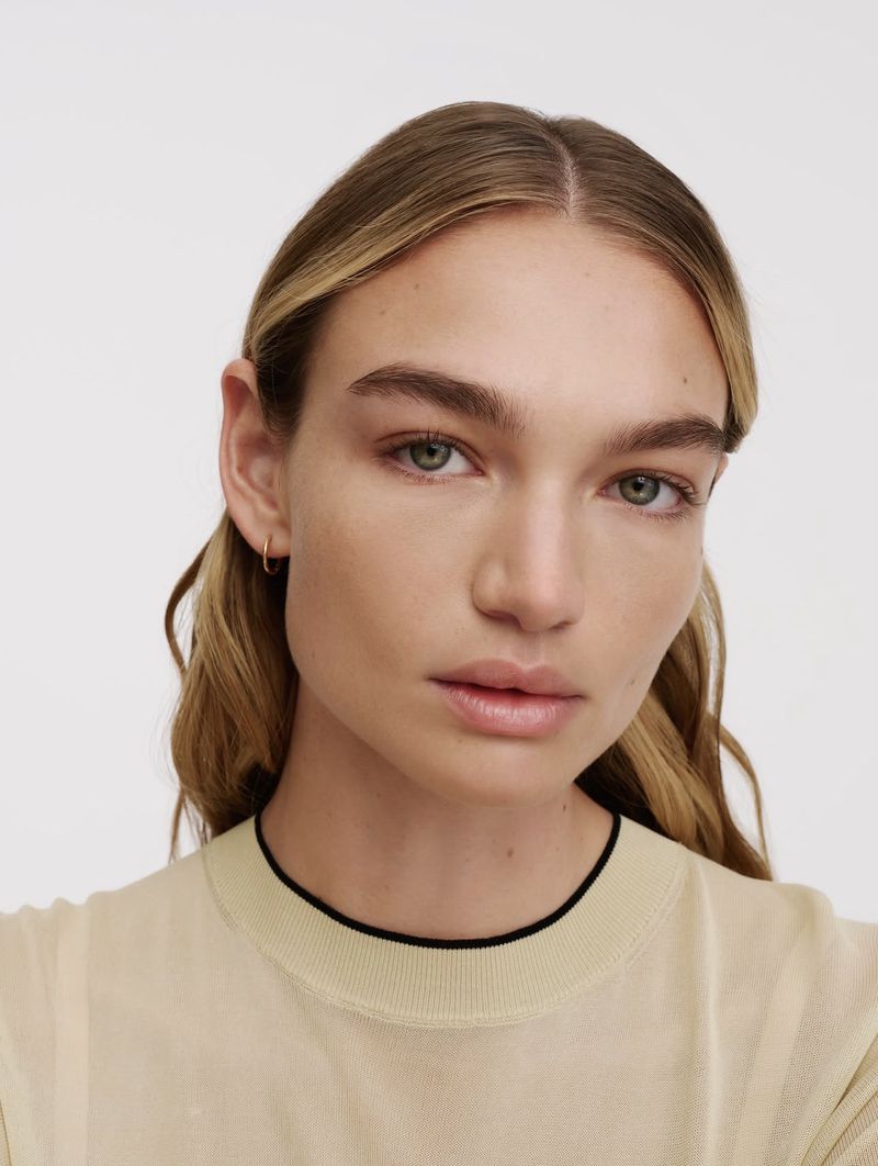 Close-up portrait of a woman with blonde highlights wearing a beige sweater with a black trim against a light background.