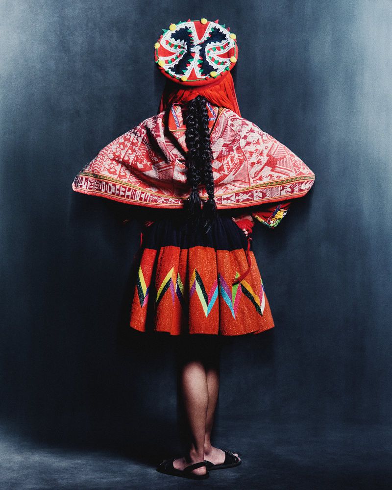 A person in traditional Andean clothing stands facing away from the camera, showcasing the intricate designs of their red and black garments.