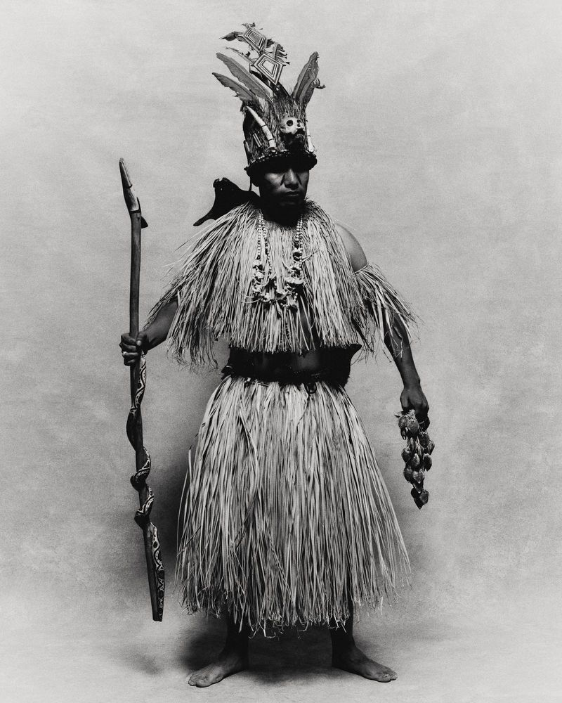 Full-length studio shot of a person in traditional dress with straw skirt, beaded necklaces, feathered headpiece, holding a staff and fruit.