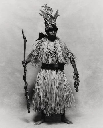 Full-length studio shot of a person in traditional dress with straw skirt, beaded necklaces, feathered headpiece, holding a staff and fruit.