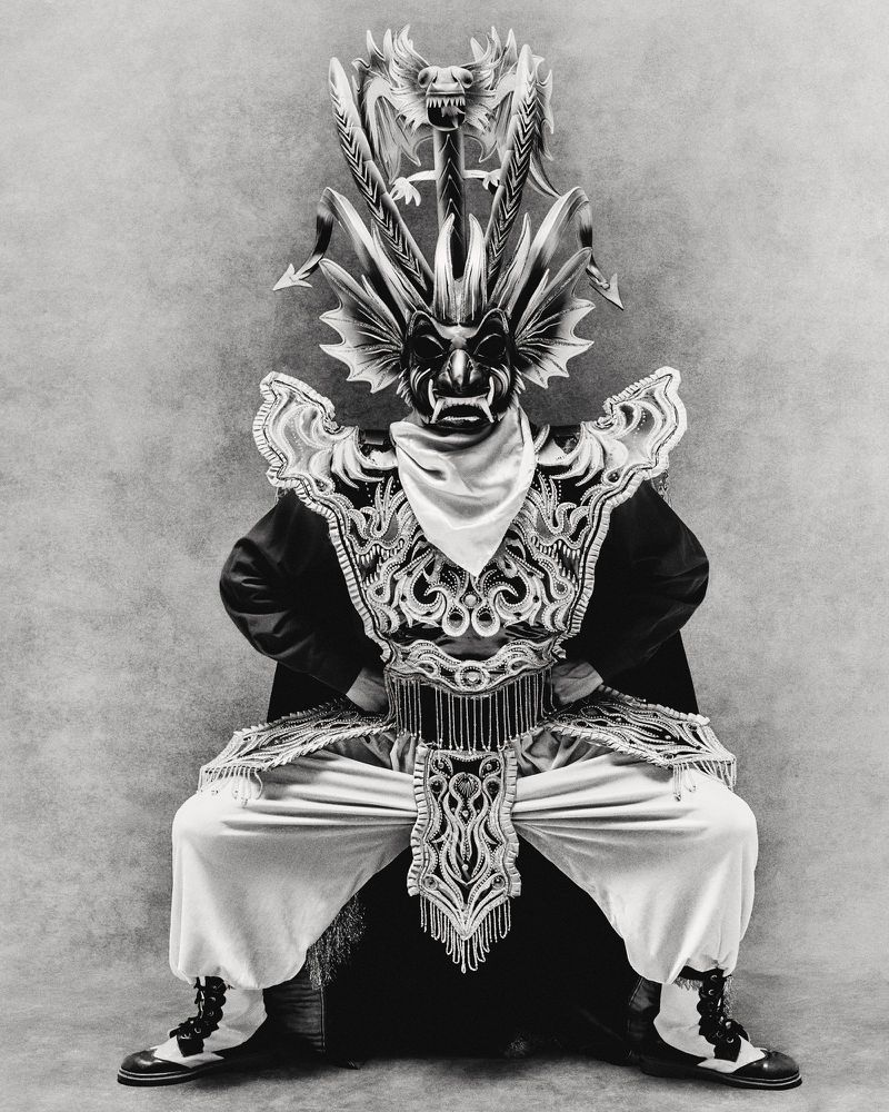 Performer in ornate costume and headdress squats with hands on hips, dramatic lighting, creating a striking black and white theatrical portrait.