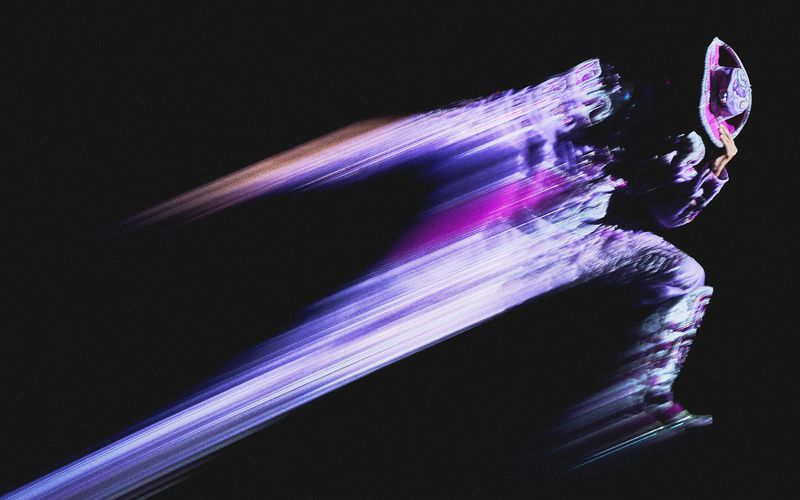 Motion blur of a person in a costume running against a black background, featuring purple and pink light streaks and a sense of speed.