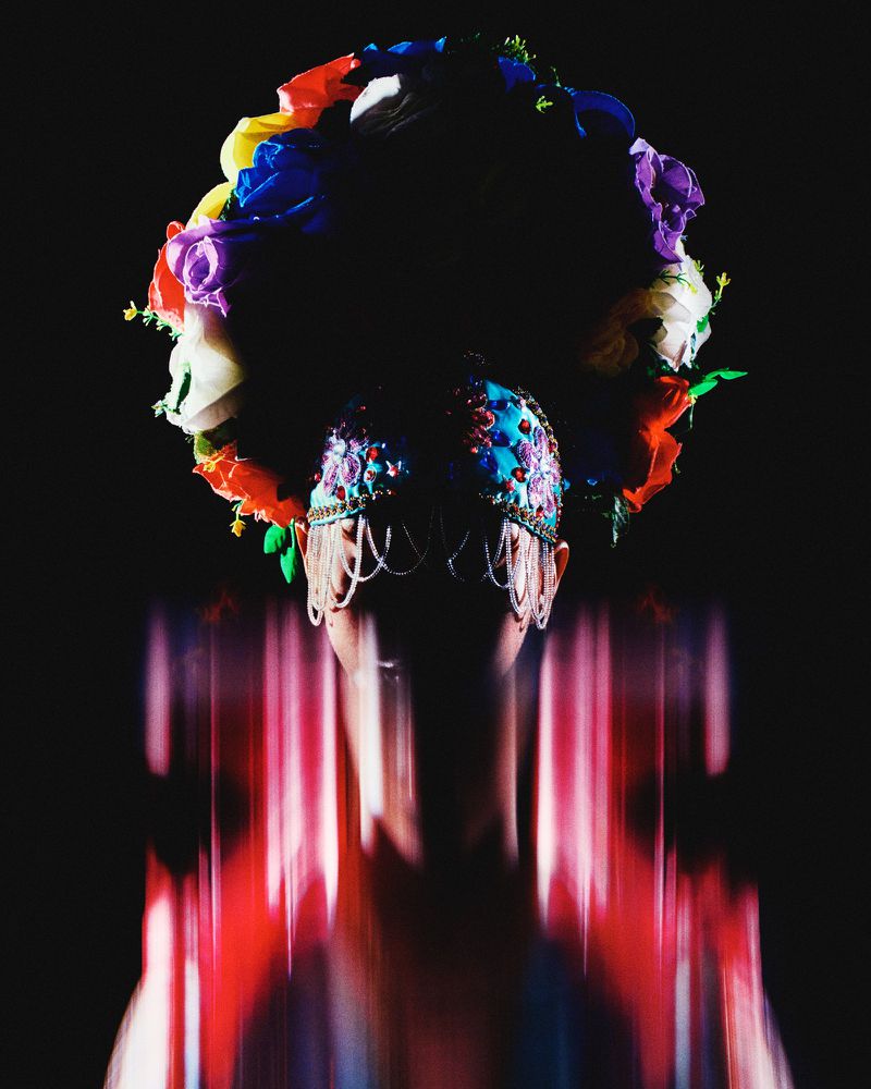 Person wearing a flower crown and ornate beaded headpiece against a dark background, blurred pink and red lines in the foreground.