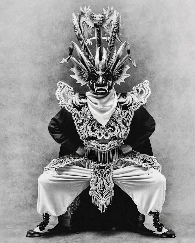 Full-length black and white portrait of a seated dancer in an elaborate dragon mask and embroidered folk costume against a textured backdrop.