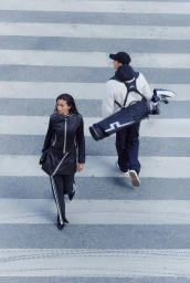 Kelly Gale in a black leather outfit and Joel Kinnaman carrying a golf bag while walking across a crosswalk.