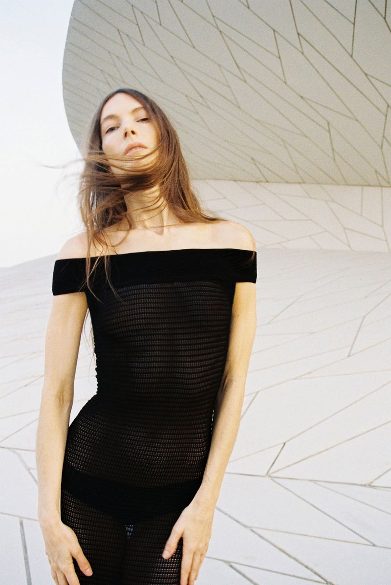 Jessica Miller in a black off-the-shoulder mesh dress standing before geometric architecture in the Mônot campaign.