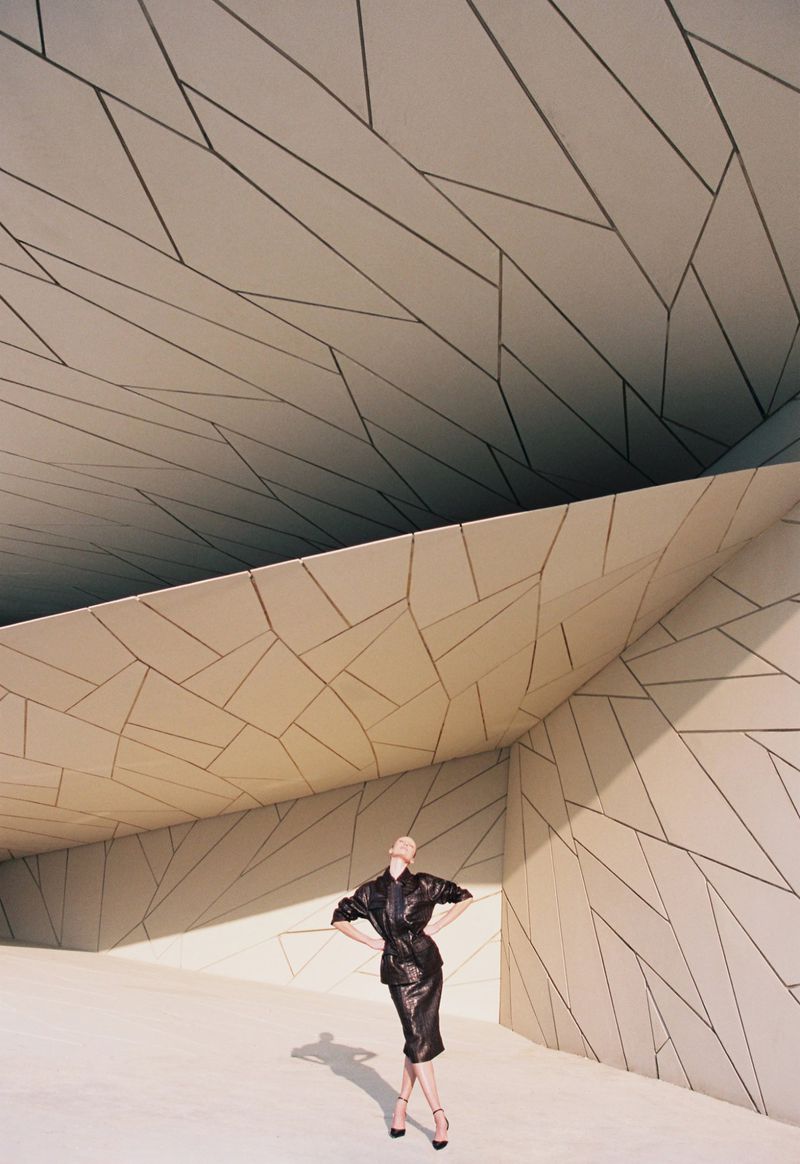 Model wearing a black leather outfit posing in a vast geometric architectural setting with angular paneled surfaces.