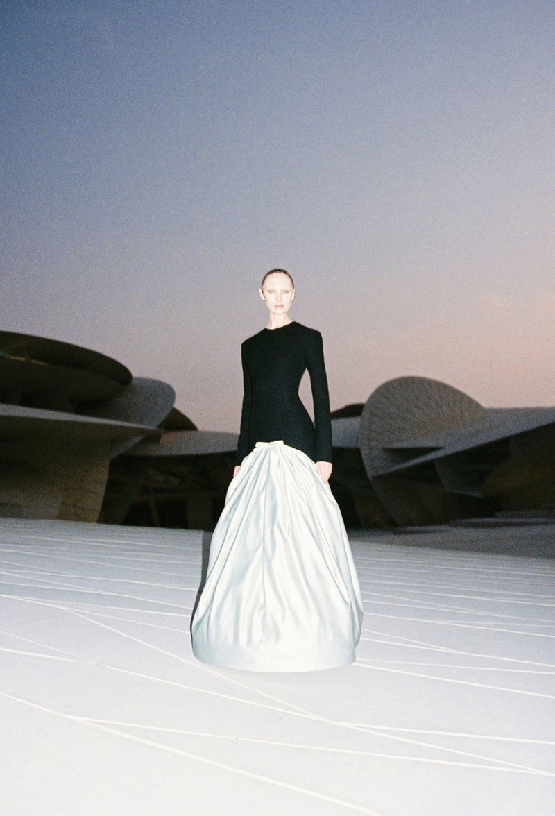 A woman stands centered, wearing a long-sleeved black bodice with a voluminous, flowing white satin skirt against a modern desert building.