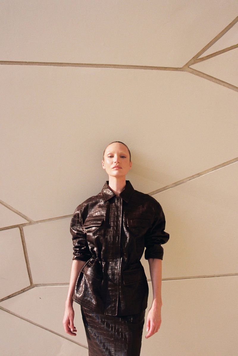 Model wearing a dark brown crocodile-embossed leather jacket and matching skirt against a geometric paneled wall.