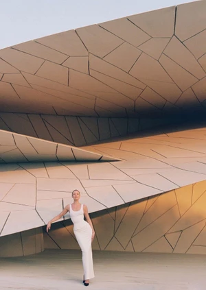 Model Candice Swanepoel wears a white maxi dress while posing against a sand-toned geometric architectural facade.
