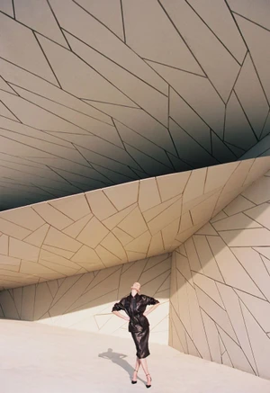Model wearing a black leather outfit posing in a vast geometric architectural setting with angular paneled surfaces.