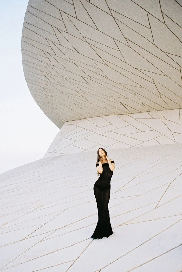 Model wearing a long black off-the-shoulder evening gown posing against a modern white geometric architectural structure.
