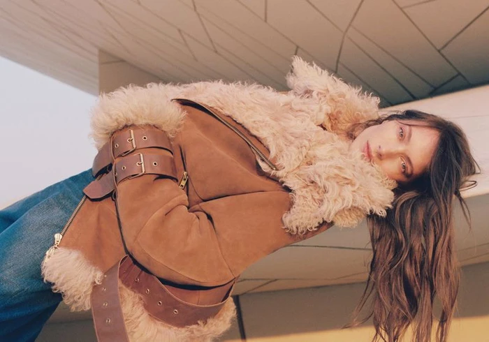 Model Jessica Miller wearing a brown shearling jacket with buckles and denim against geometric architecture for Mônot.