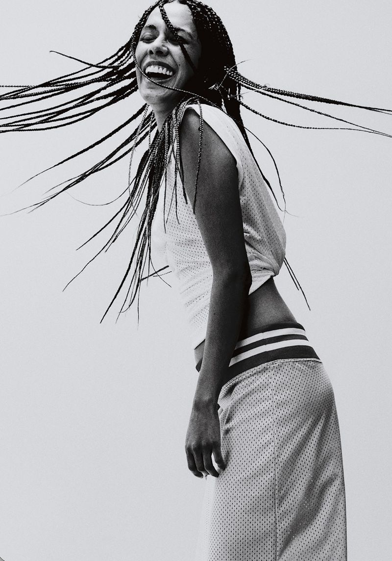 A smiling woman in a white perforated athletic mesh top and skirt poses in black and white as her braided hair whips through the air.