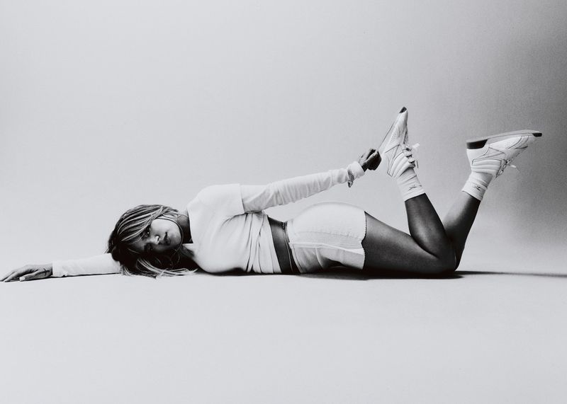 Karol G lying on her side in white athletic apparel and sneakers against a seamless studio backdrop.