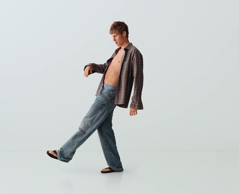 Man posing in an open plaid button-up shirt and baggy blue denim jeans against a seamless white studio background.