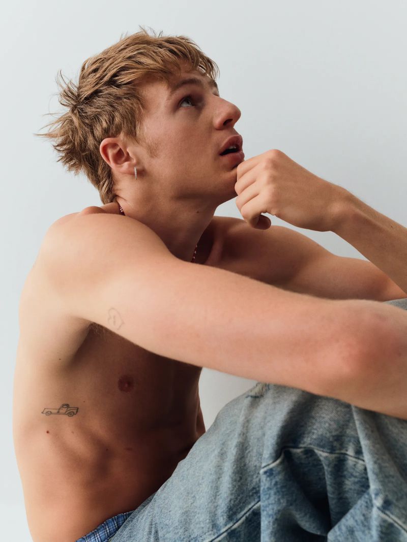 Young man with messy blonde hair and a small car tattoo on his side wearing light blue denim jeans against a plain white background.