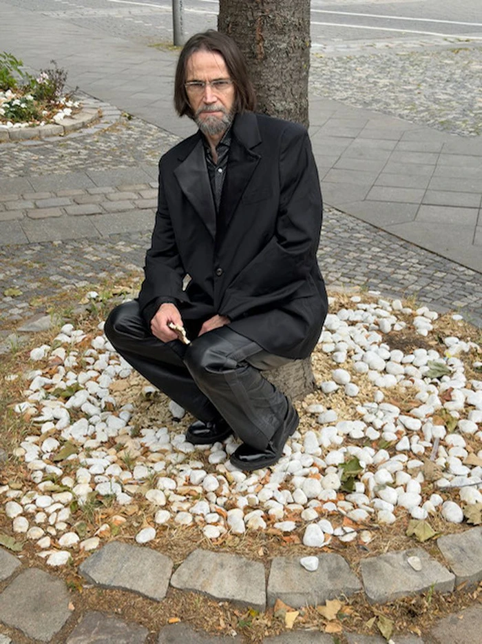 Karl Heinz Hirschberger crouching by a tree in a black blazer and leather trousers on a cobblestone sidewalk.
